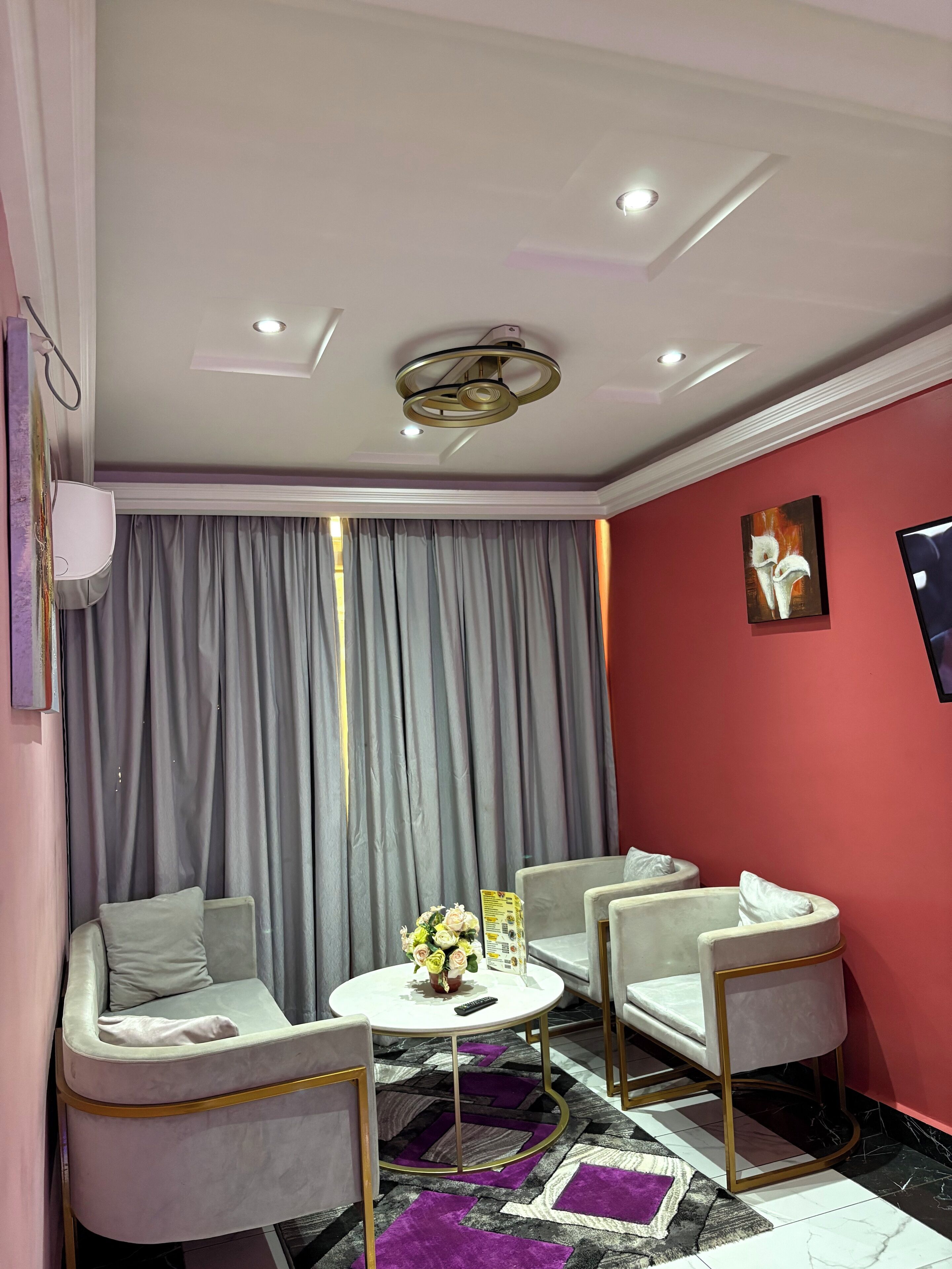 Un salon moderne avec une table basse ronde blanche, deux fauteuils blancs, un rideau gris, un mur d’accent rouge et un luminaire au plafond.