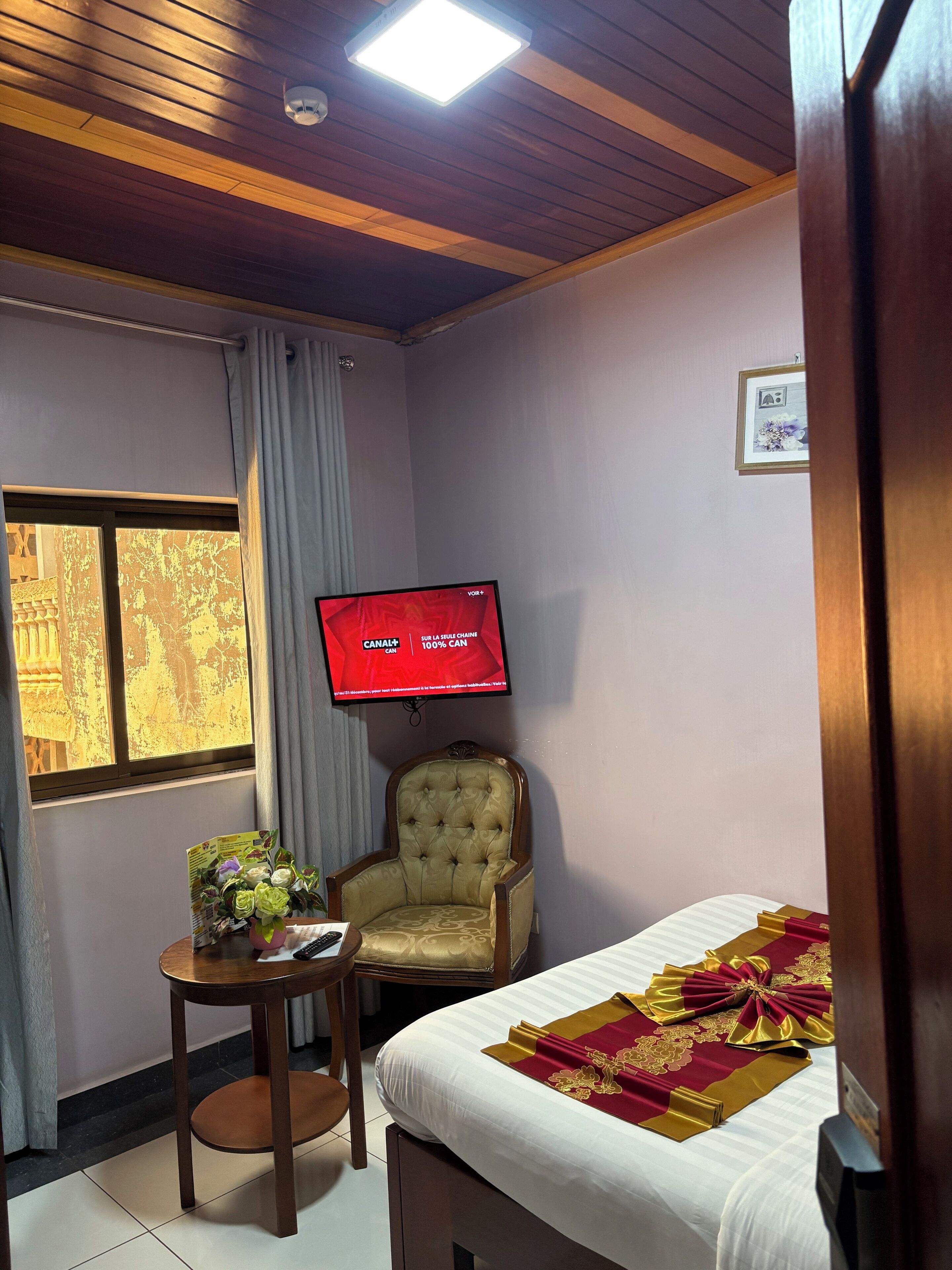 Ein Hotelzimmer mit Bett, Holzboden, Fernseher, Sessel, kleinem Tisch mit Blumenstrauß und Fenster mit Vorhängen.