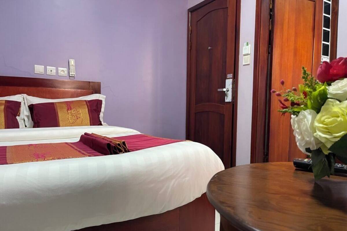 A hotel room with a bed, a wooden door, a round table, and a vase of flowers.