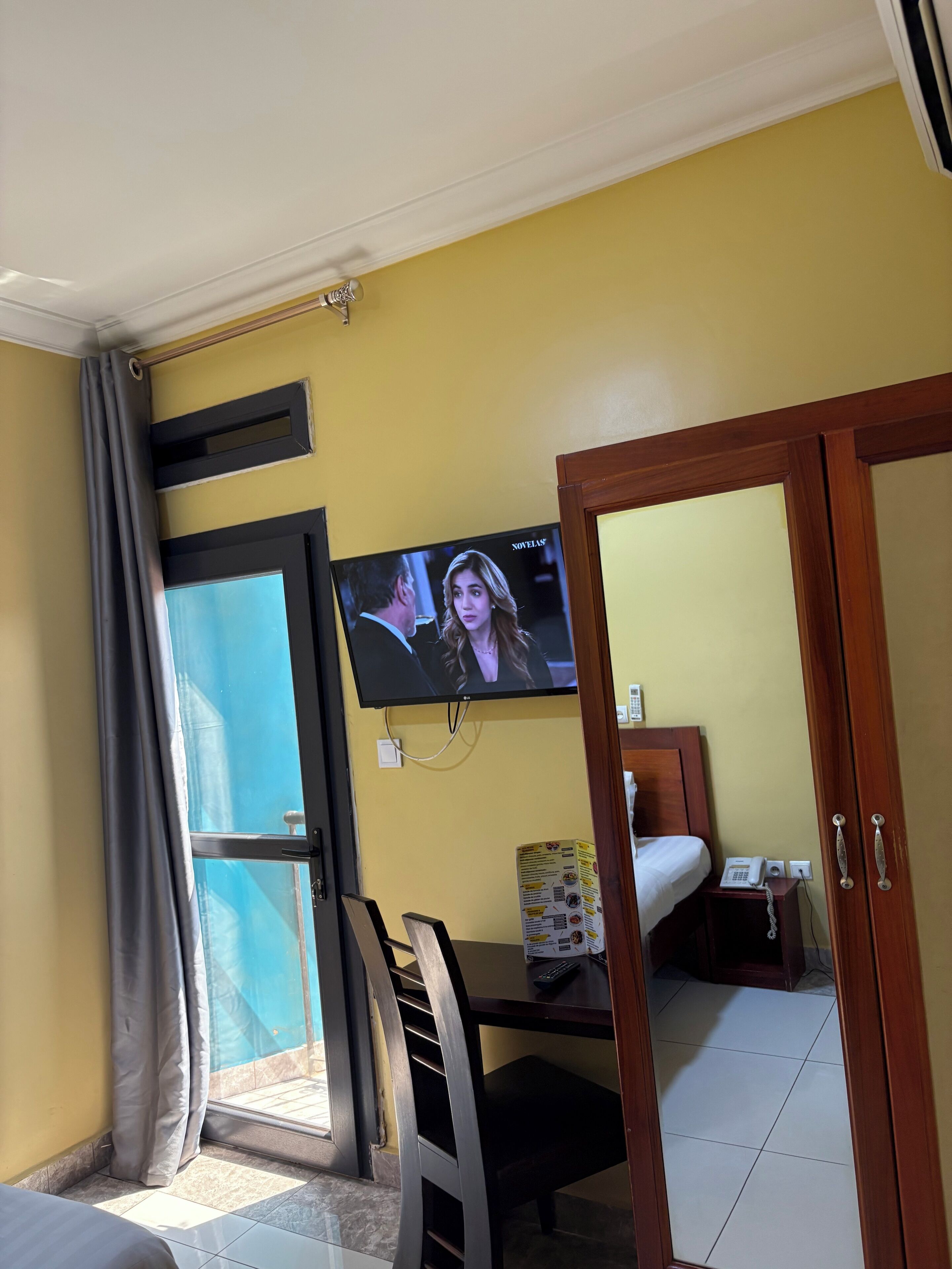 Ein Hotelzimmer mit an der Wand befestigtem Fernseher, einem Bett mit weißer Bettdecke, einem kleinen Nachttisch mit Telefon und Fernbedienung sowie einem Fenster mit Vorhängen.