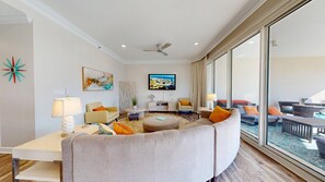 A modern living room with a curved sofa, a flat-screen TV, and a large window offering a view of the outdoors.