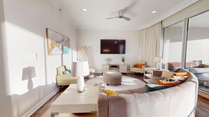 Una sala de estar moderna con un sofá, una mesa de centro y sillones. Hay un televisor de pantalla plana montado en la pared, un ventilador de techo y un ventanal con cortinas.