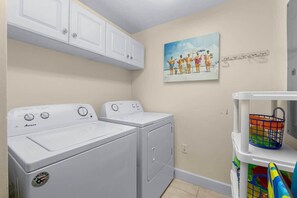 A laundry room with two white Amana washers and dryers, a painting on the wall, and a shelf with baskets.