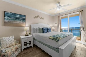 A bedroom with a bed, nightstands, a ceiling fan, a window with a view of the ocean, and a beach-themed pillow.