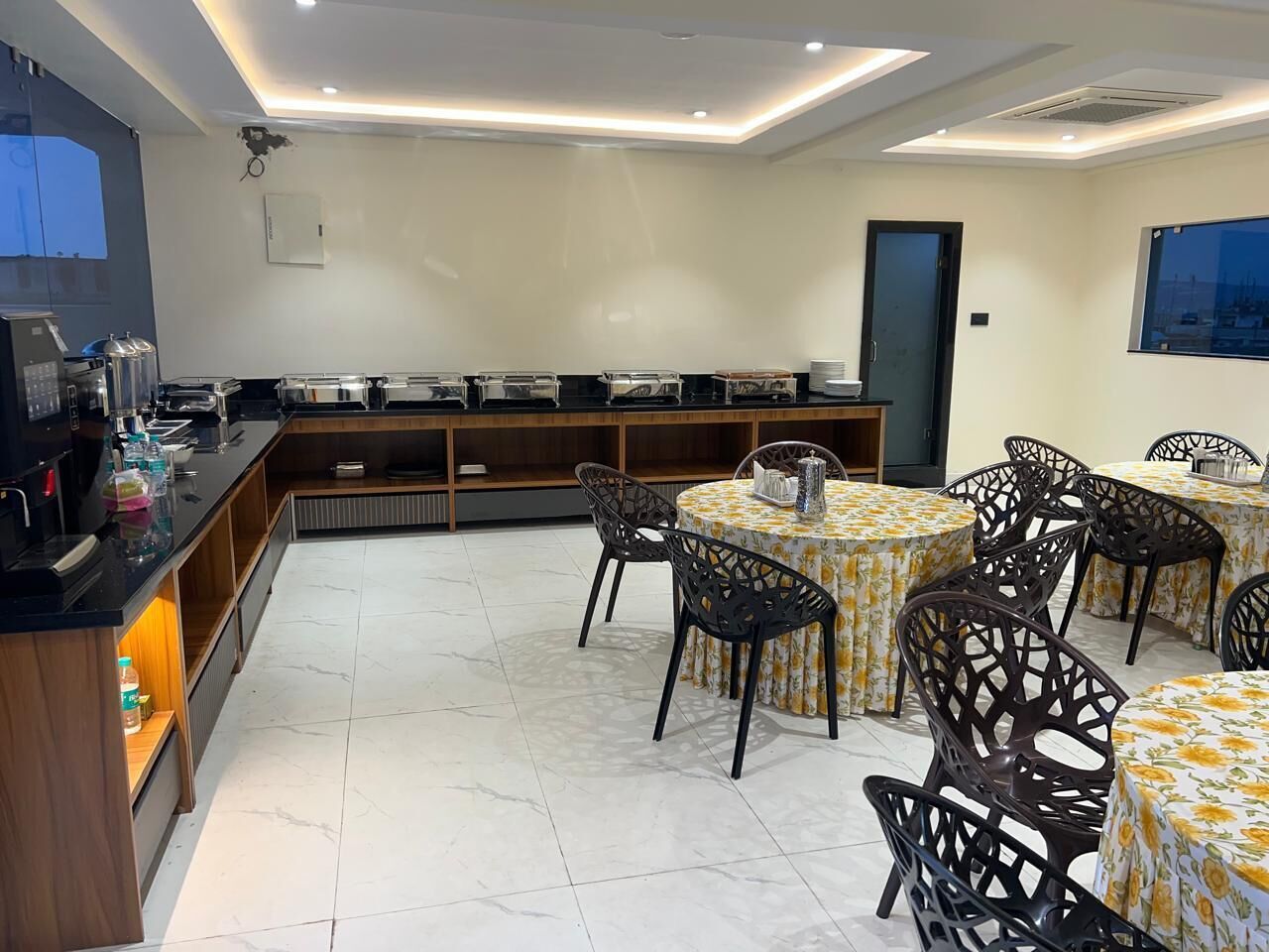 Un espace repas avec des tables rondes recouvertes de nappes à motifs floraux, des chaises noires et un buffet où sont disposés des récipients contenant de la nourriture.