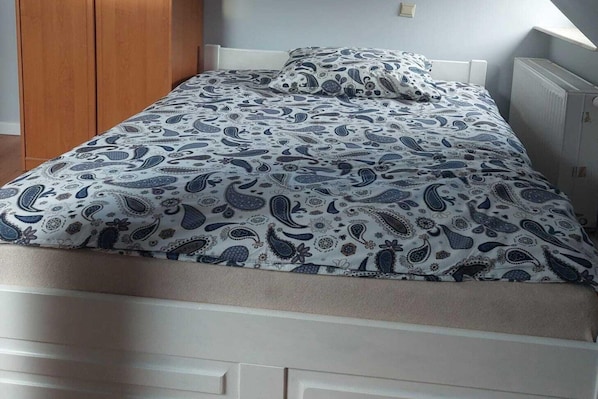 A bed with a patterned bedspread and pillows, a wooden wardrobe, and a radiator.