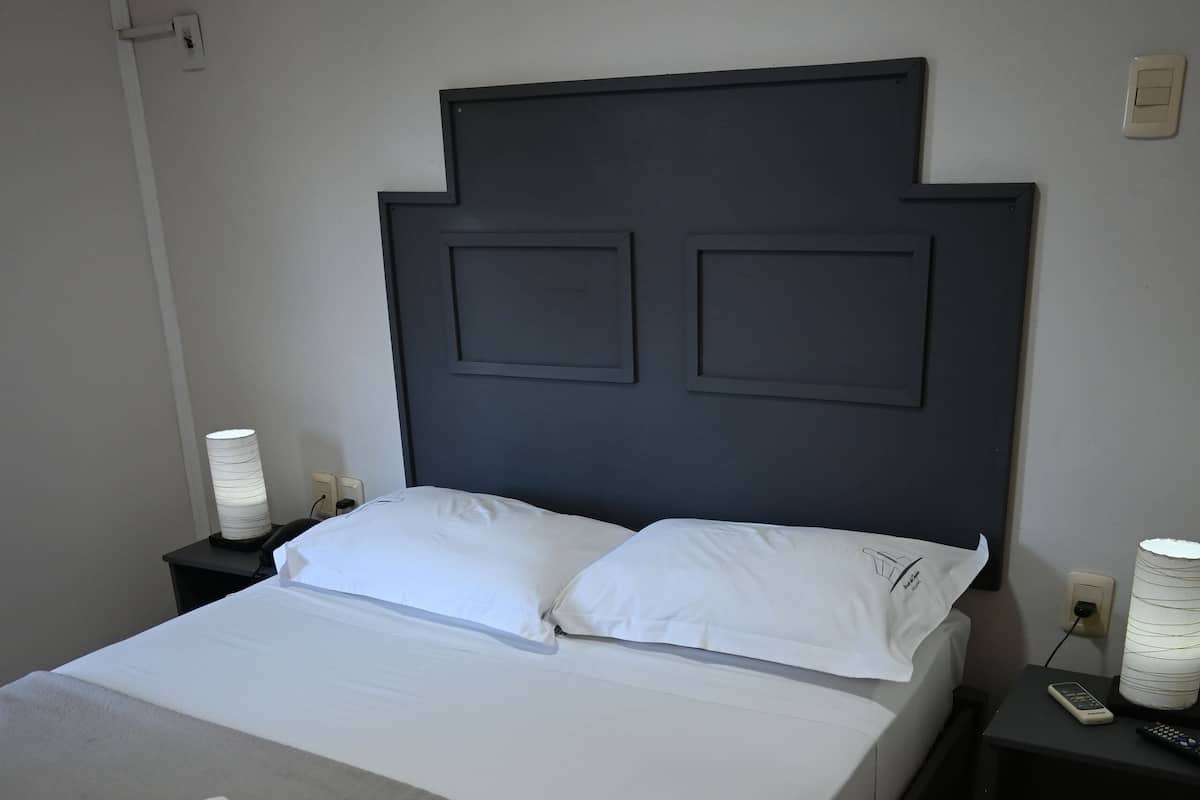 A neatly made bed with white linens, a dark headboard, and a bedside table with a lamp and a remote control.