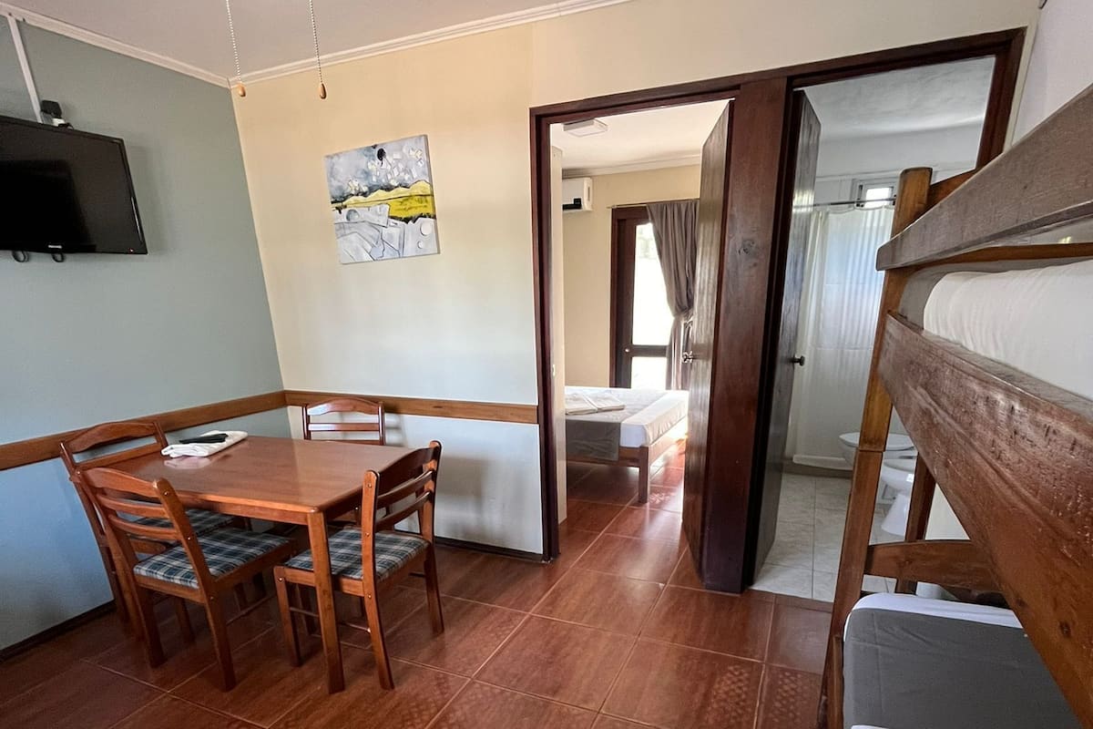 A room with a wooden dining table and chairs, a ceiling fan, a bed, and a shower area.