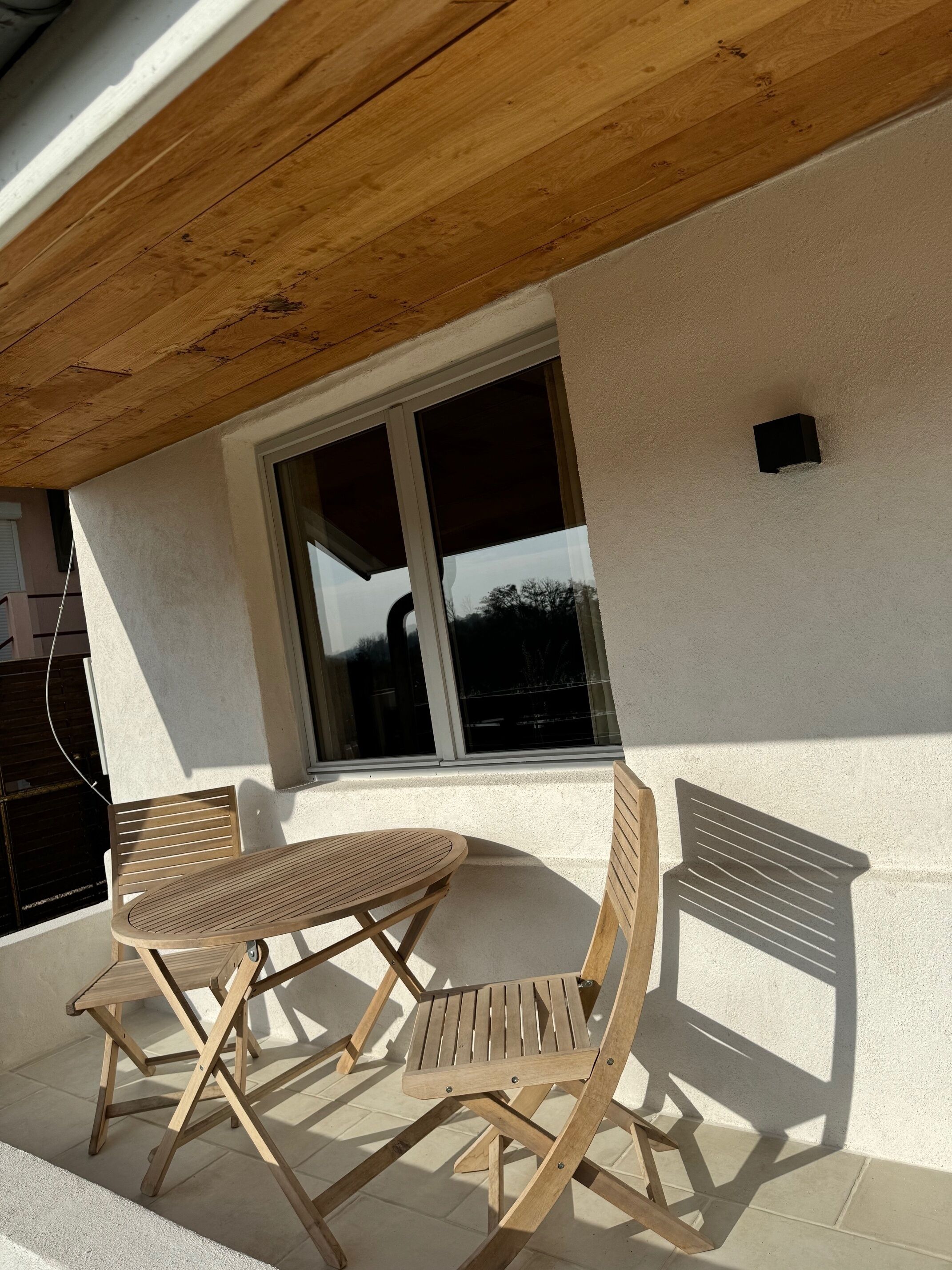 Une table en bois et deux chaises sont placées à l’extérieur d’un bâtiment à la façade blanche et doté d’une grande fenêtre.