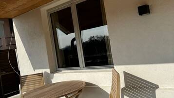 Une table en bois et deux chaises sont placées à l’extérieur d’un bâtiment à la façade blanche et doté d’une grande fenêtre.