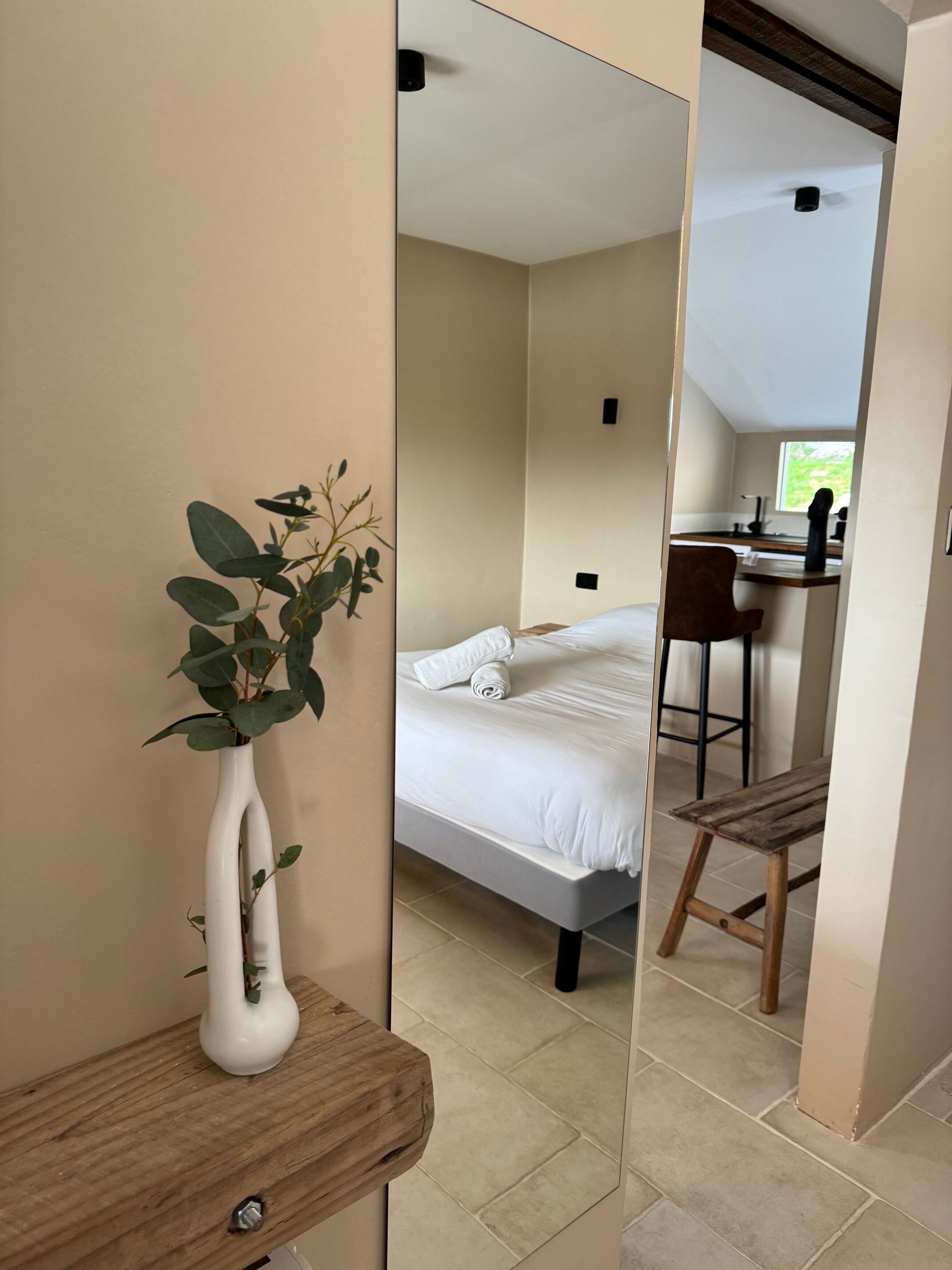 Een slaapkamer met een bed, een houten stoel, een witte vaas met een plant, en uitzicht op een keuken door een deuropening.