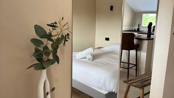 Een slaapkamer met een bed, een houten stoel, een witte vaas met een plant, en uitzicht op een keuken door een deuropening.