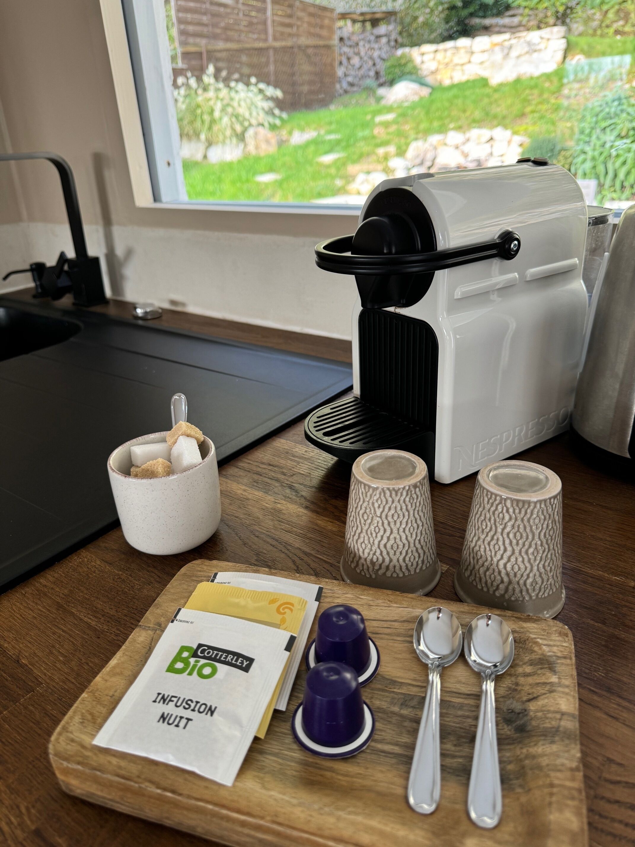 Een keukenblad met een koffieapparaat, een mok, een pak koffiecups en een set lepels.