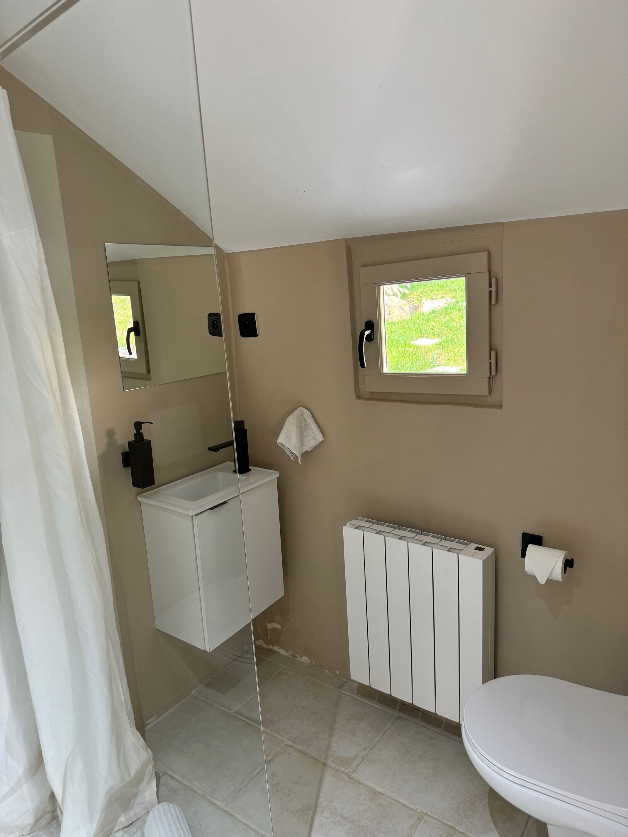 A bathroom with a white sink, a toilet, a radiator, a window with a view of greenery, and a mirror.
