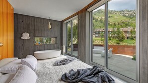 A modern bedroom with a large bed, a bathtub, and a view of a green landscape through large windows. - Architectural Gem by InvitedHome | Ski-In, Sauna, Patio, Firepit, Unique (Alta)