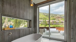 A modern bathroom with a freestanding bathtub, a large window, and a view of a hillside and buildings. - Architectural Gem by InvitedHome | Ski-In, Sauna, Patio, Firepit, Unique (Alta)