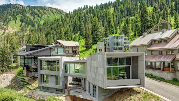 A modern multi-story building with large windows and a stone facade, surrounded by lush greenery and mountains. - Architectural Gem by InvitedHome | Ski-In, Sauna, Patio, Firepit, Unique (Alta)