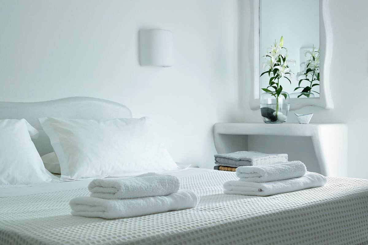 Un lit bien fait, avec du linge de lit et des serviettes blanches, une table de chevet ornée d’un vase de fleurs et une fenêtre donnant sur un paysage verdoyant.