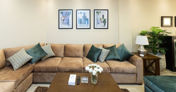 A living room with a brown sectional sofa, a coffee table with a remote control and a vase of flowers, and framed artwork on the wall. - Prime Select Lotus New Cairo (New Cairo)