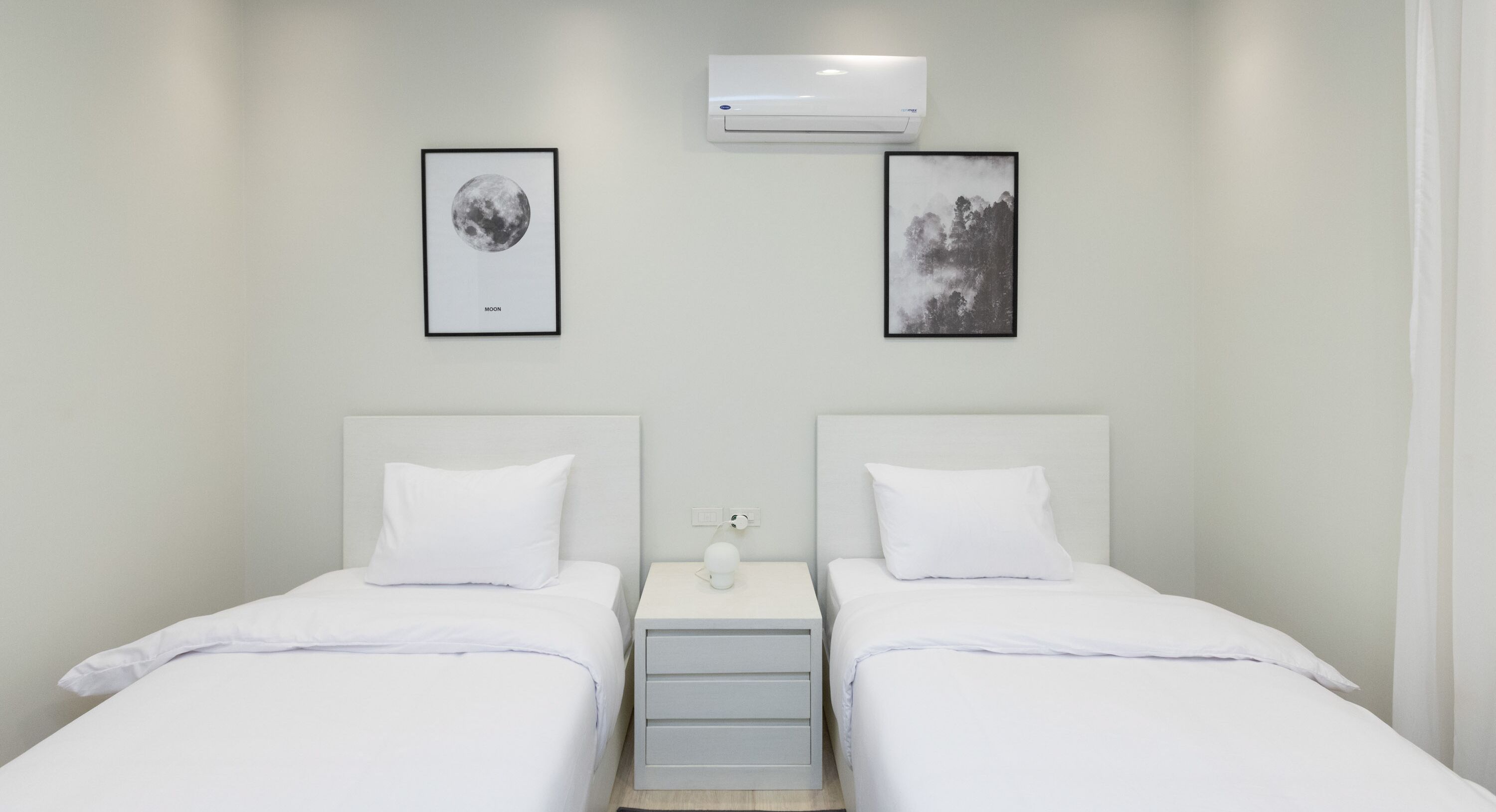 Une chambre d’hôtel avec deux lits, du linge de lit blanc, une table de chevet avec une lampe et deux tableaux encadrés au mur.