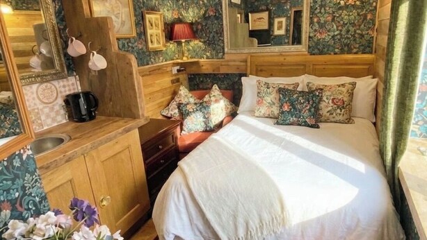 Ein Schlafzimmer mit Holzkopfstütze, blumigem Wandpapier, einem großen Spiegel, einem Bett mit vielen Kissen und einem Nachttisch mit Lampe.