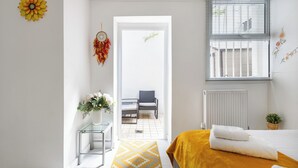 A bedroom with a bed, a window with blinds, a radiator, a chair, a small table, and a decorative wall hanging.