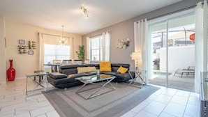 Ein modernes Wohnzimmer mit einem Ecksofa, einem Glastisch und einer großen Schiebeglastür, die zu einem Außenbereich führt.
