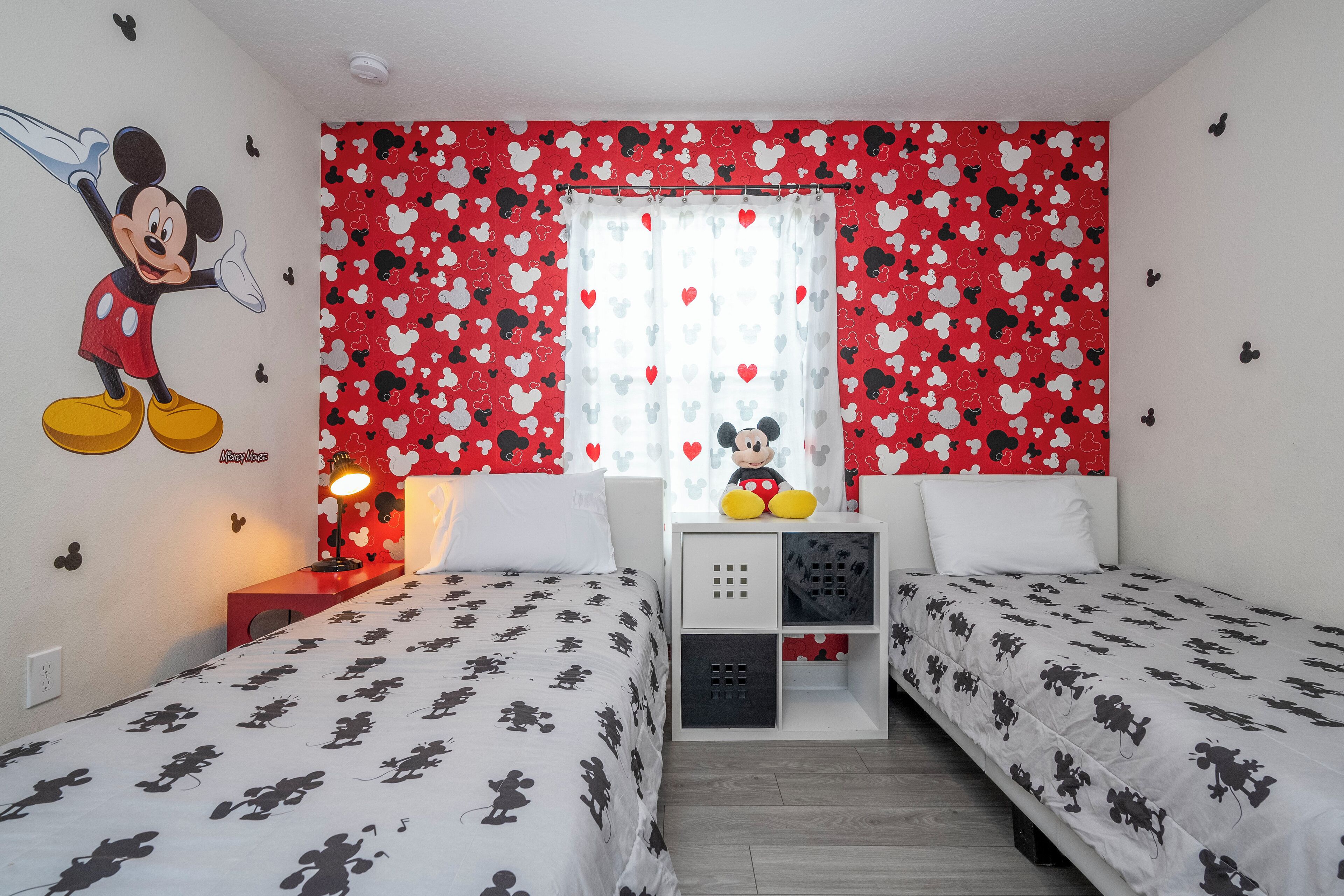 A room with two beds, a Mickey Mouse mural, a Mickey Mouse plush toy, and a window with heart-patterned curtains.