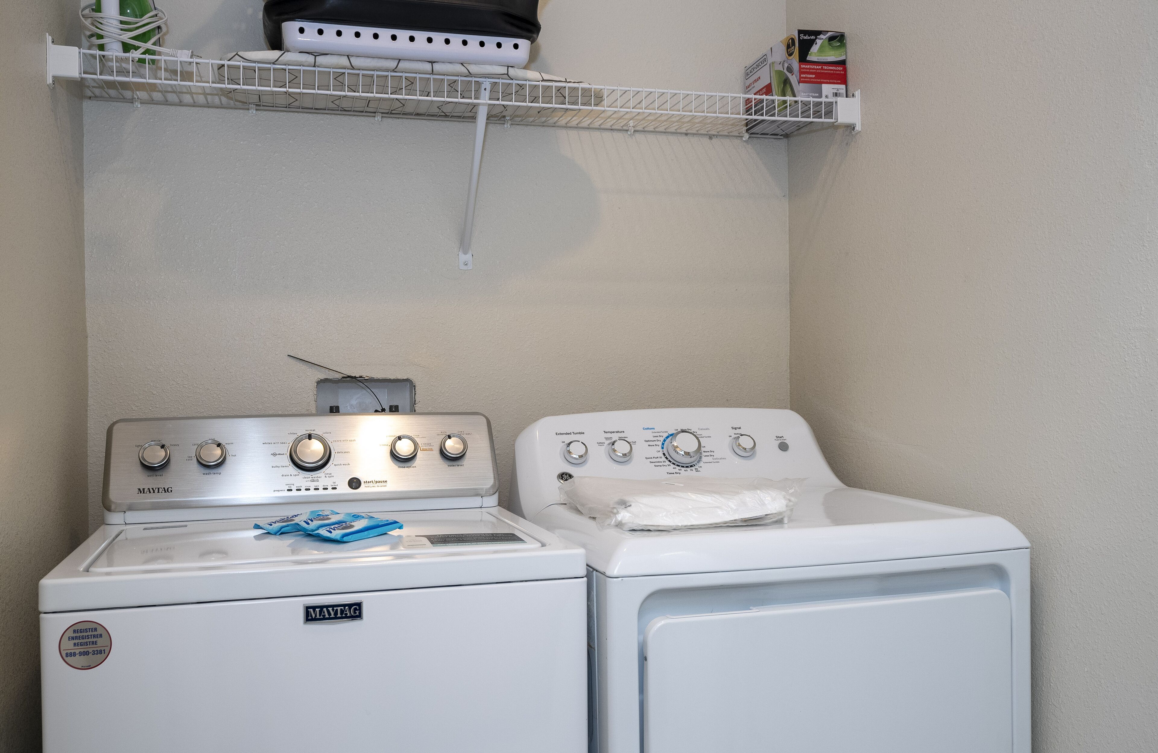 A laundry area with a front-loading washer and dryer, a shelf above with a box and a green item, and a white wall.
