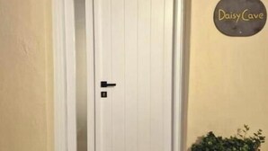 Una puerta blanca con un picaporte negro y un pequeño ventanal encima. A la derecha de la puerta hay una planta en maceta.