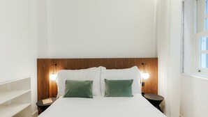 A neatly made bed with white linens and two green pillows, flanked by two bedside tables with lamps, in a room with white walls and a wooden headboard. - Feel Porto Cl Rigos Charm Flat 2 3 (Porto)