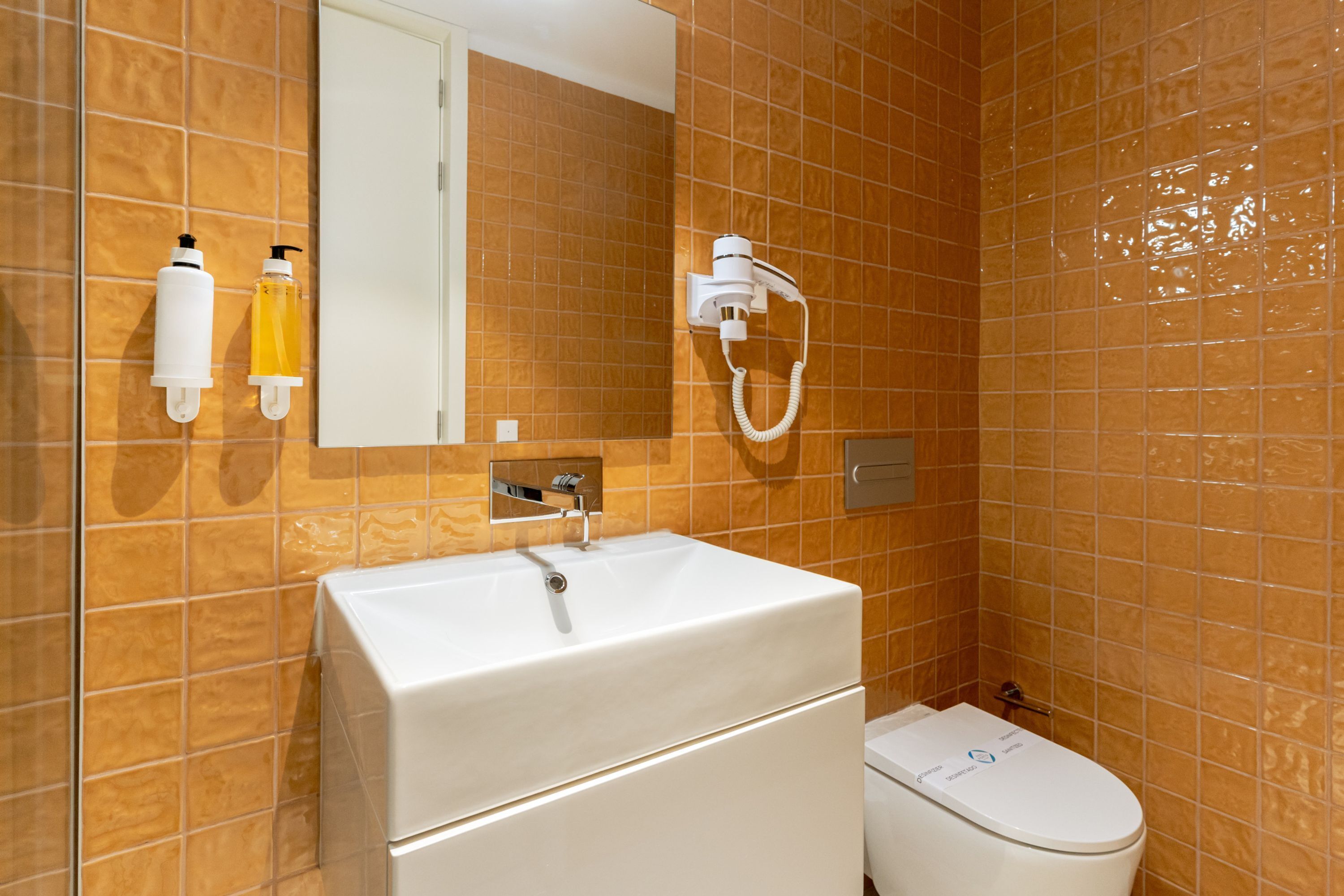 Une salle de bain avec un lavabo blanc, un miroir et des toilettes. On y trouve deux flacons de savon liquide et un sèche-cheveux fixé au mur.