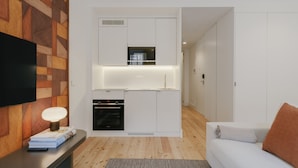 Cozinha moderna com armários brancos, forno embutido e micro-ondas. Há uma televisão de tela plana na parede, uma parede de destaque em madeira e um tapete cinza no chão.