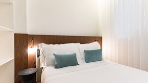 Quarto de hotel bem organizado, com cabeceira de madeira, roupa de cama branca e uma pequena mesa de apoio.