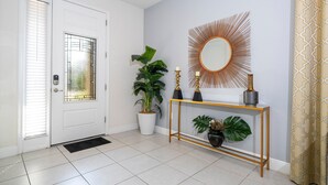 Ein heller, moderner Eingangsbereich mit einer weißen Tür, einem Blumentopf, einem Konsolentisch mit Dekoration und einem Spiegel in Form einer Sonne an der Wand.