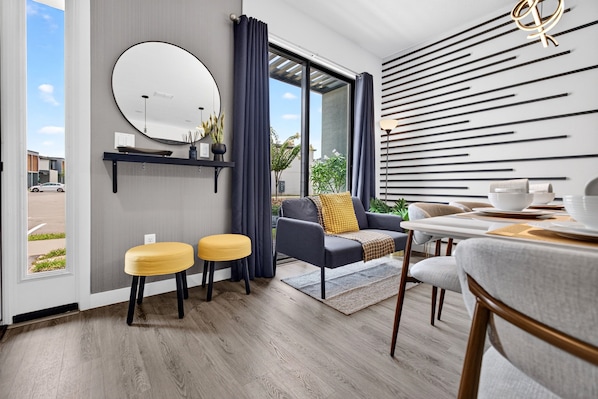 Un intérieur moderne comprenant un coin repas, un canapé avec un coussin jaune, un miroir rond et une vue à travers une porte vitrée.