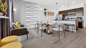 A modern kitchen with a central island, a dining area with a round table and chairs, and a living area with a sofa and yellow ottoman.