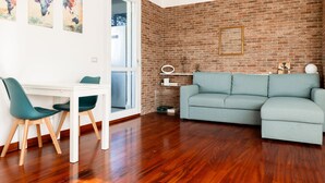 A living room with a brick wall, a light blue sofa, a white table with green chairs, and a small shelf with decorative items.