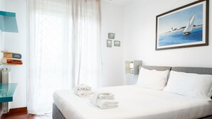A neatly made bed with white linens, a glass shelf with books, a painting of a sailboat, and a window with sheer curtains.