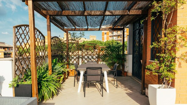 A covered patio with a table and chairs, surrounded by potted plants and a wooden fence.