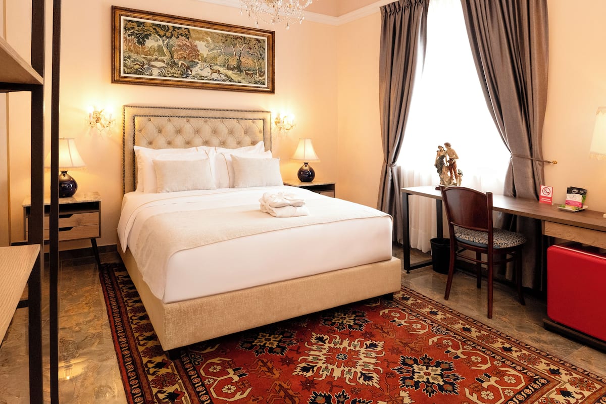 Une chambre d’hôtel avec un grand lit, un bureau, une chaise, un lustre, un tableau et une armoire rouge.
