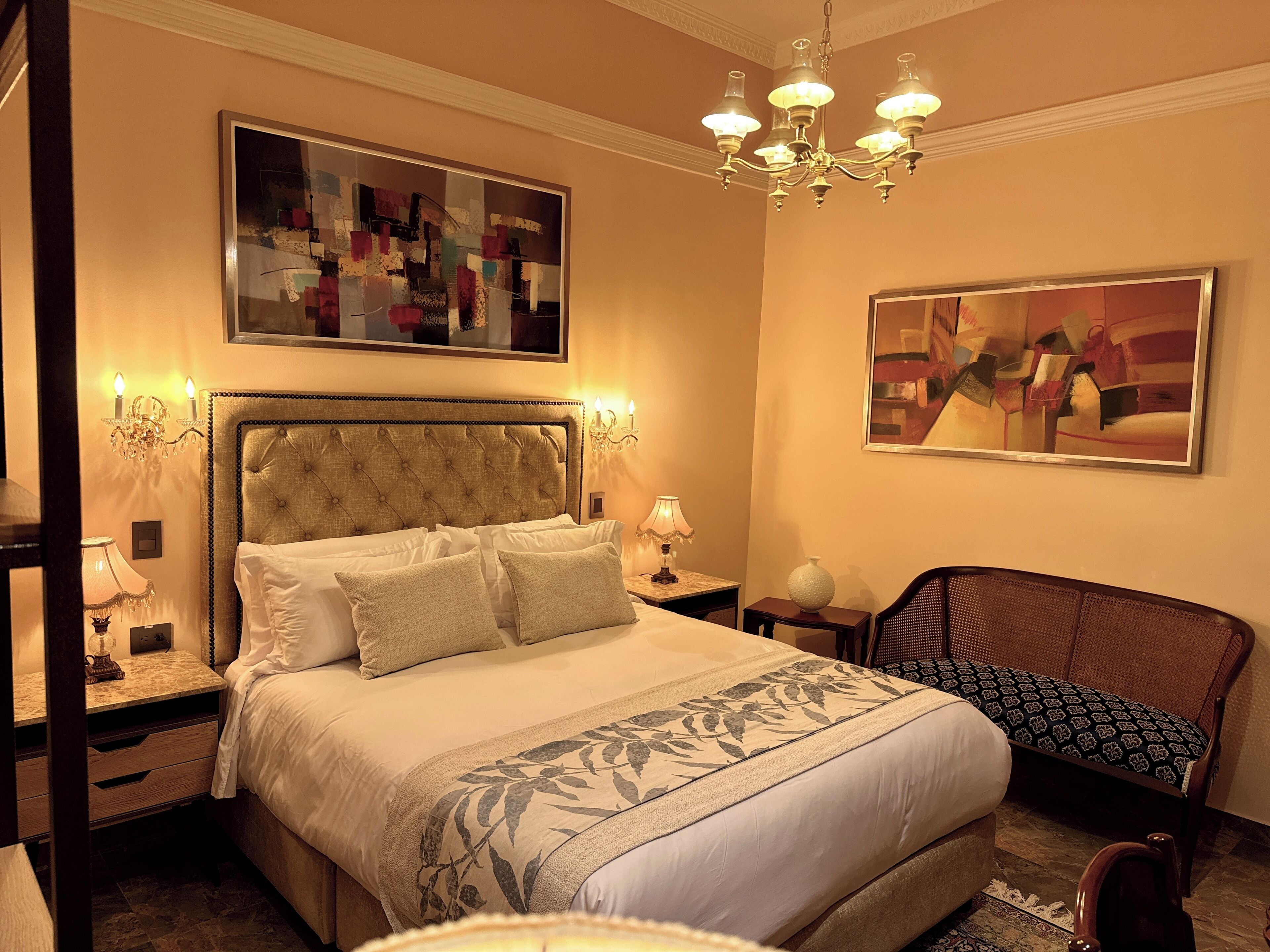 A hotel room with a large bed, a chandelier, two paintings, and a chair.
