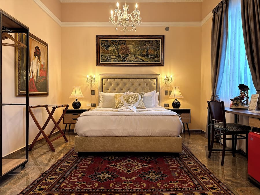 A spacious bedroom with a large bed, two bedside tables with lamps, a chandelier, a painting above the bed, and a view of the city through a window.