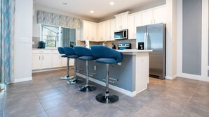 A modern kitchen with white cabinets, a stainless steel refrigerator, a microwave, and a bar area with blue bar stools.