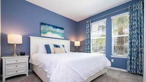 A bedroom with a white bed, two bedside tables with lamps, a blue wall, and a window with patterned curtains.