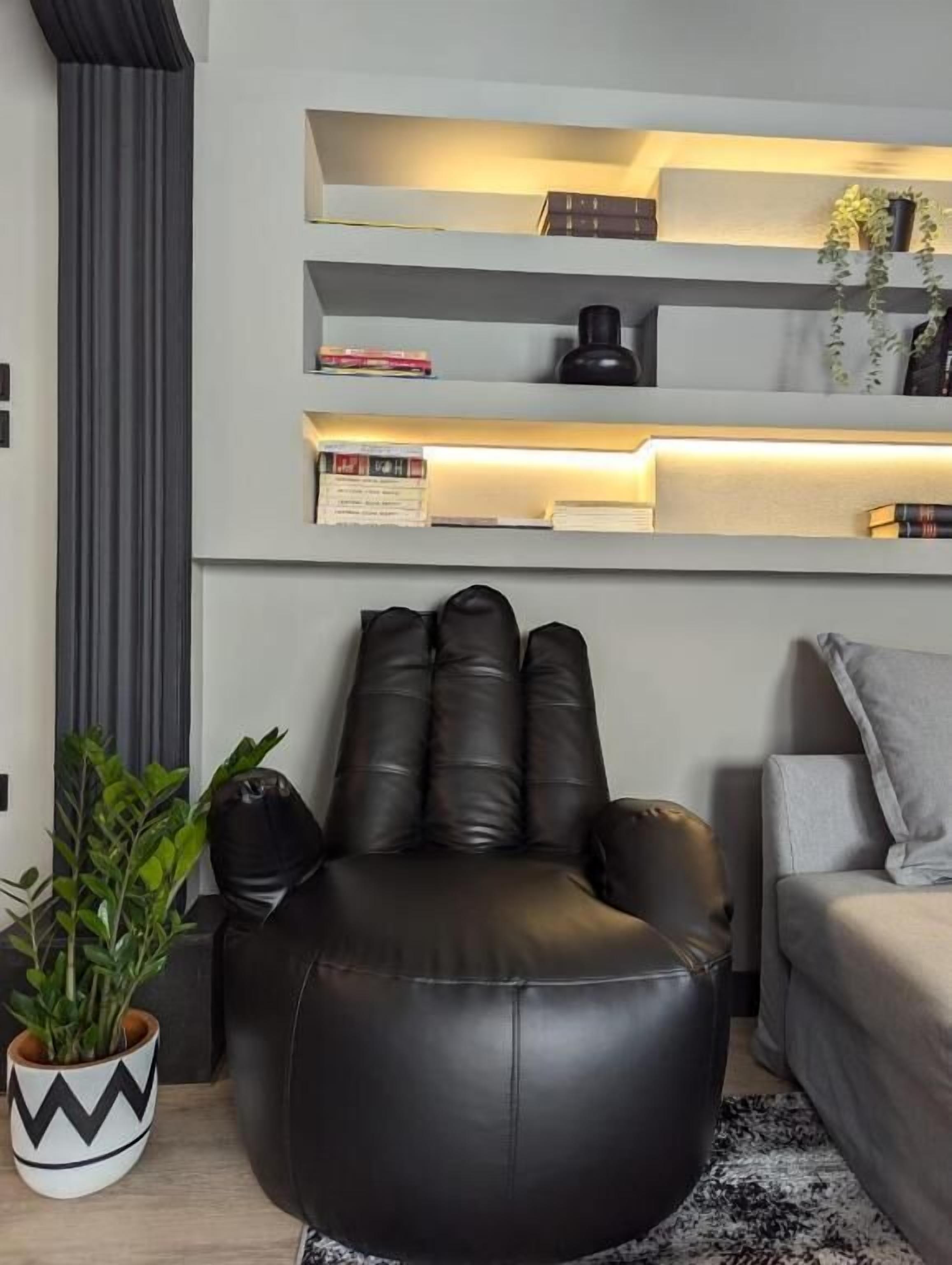 A modern living room with a black leather ottoman, a grey sofa, and a shelving unit with books and decorative items.