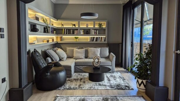 Una sala moderna con un sofá, un sillón de orejero negro y una mesa de centro. Se observa una estantería con libros y objetos decorativos, además de un ventanal con cortinas.