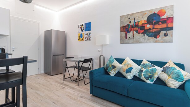 Una sala moderna con un sofá azul verdoso, una mesa de comedor con sillas, un refrigerador y arte abstracto en las paredes.