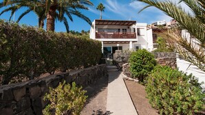 A modern two-story house with a stone pathway leading to the entrance, surrounded by lush greenery and palm trees.