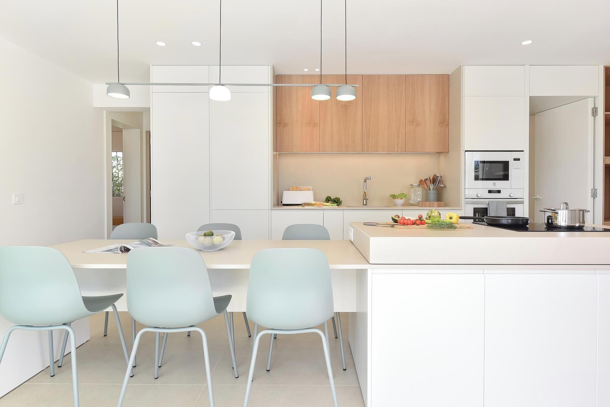 Een moderne keuken met witte kasten, een centraal eiland met een magnetron en kookplaat, en lichtblauwe stoelen rond een tafel.